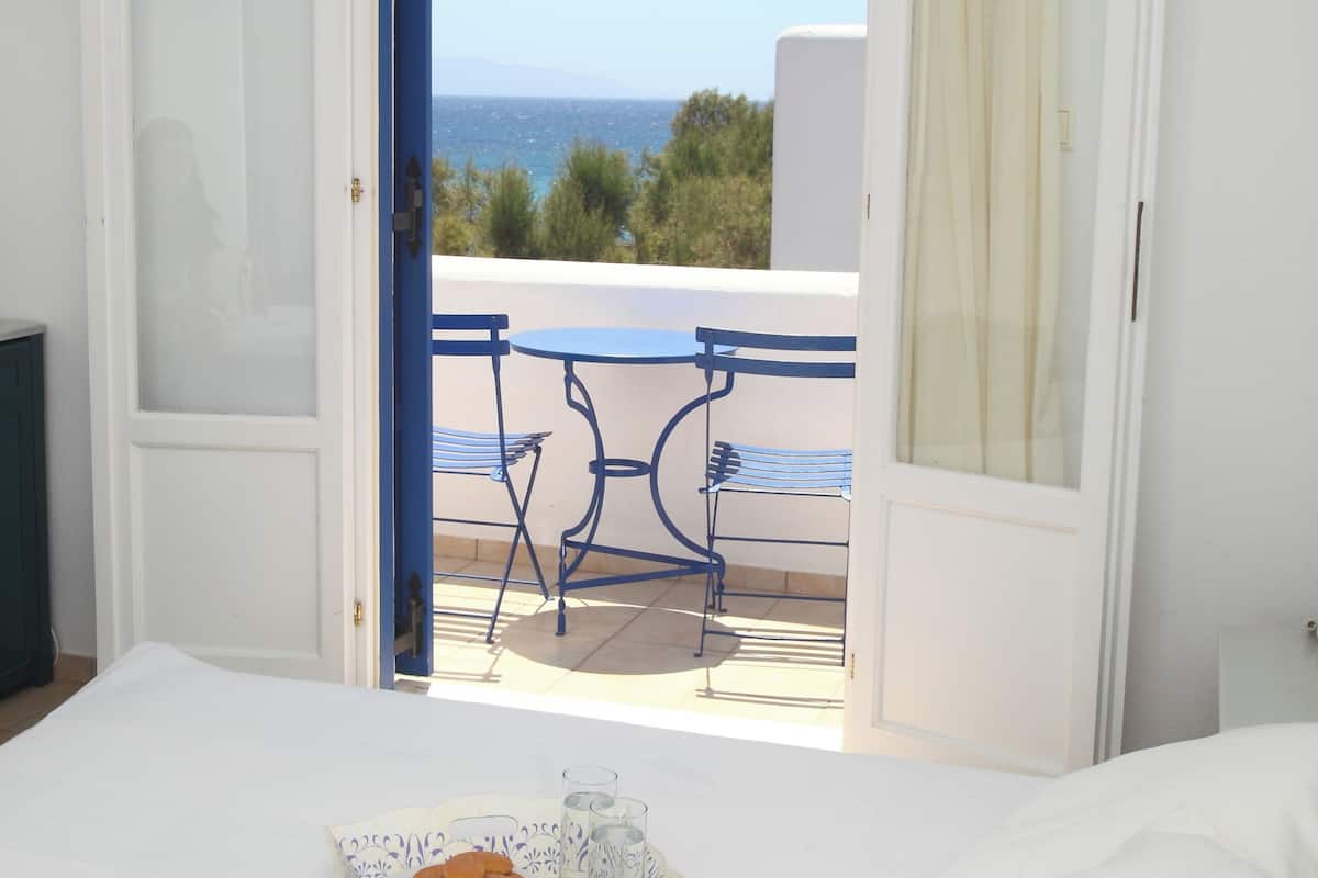 Puerta abierta de un dormitorio que da a un balcón con mesa y sillas, una bandeja con galletas y tazas de café, y vista al mar.