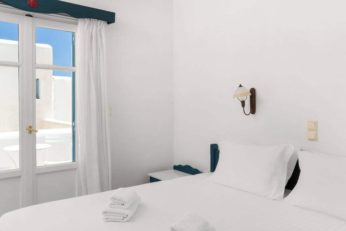 Habitación de hotel con cama, mesita de noche, lámpara y vista al exterior.