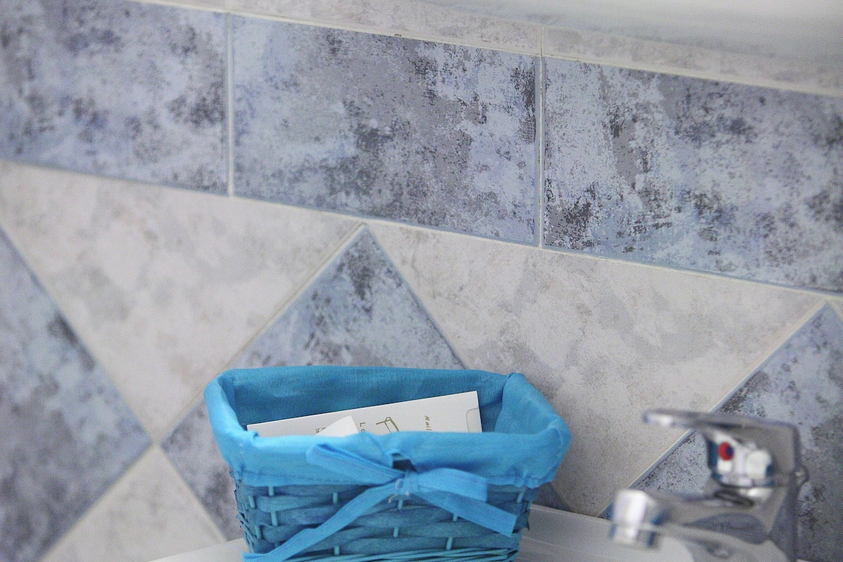 Un baño con un lavamanos blanco, una cesta azul y una pared con azulejos.
