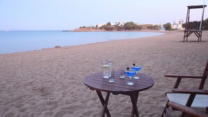 Una mesa de madera con bebidas y vasos sobre una playa arenosa, con vista al mar y edificios al fondo.