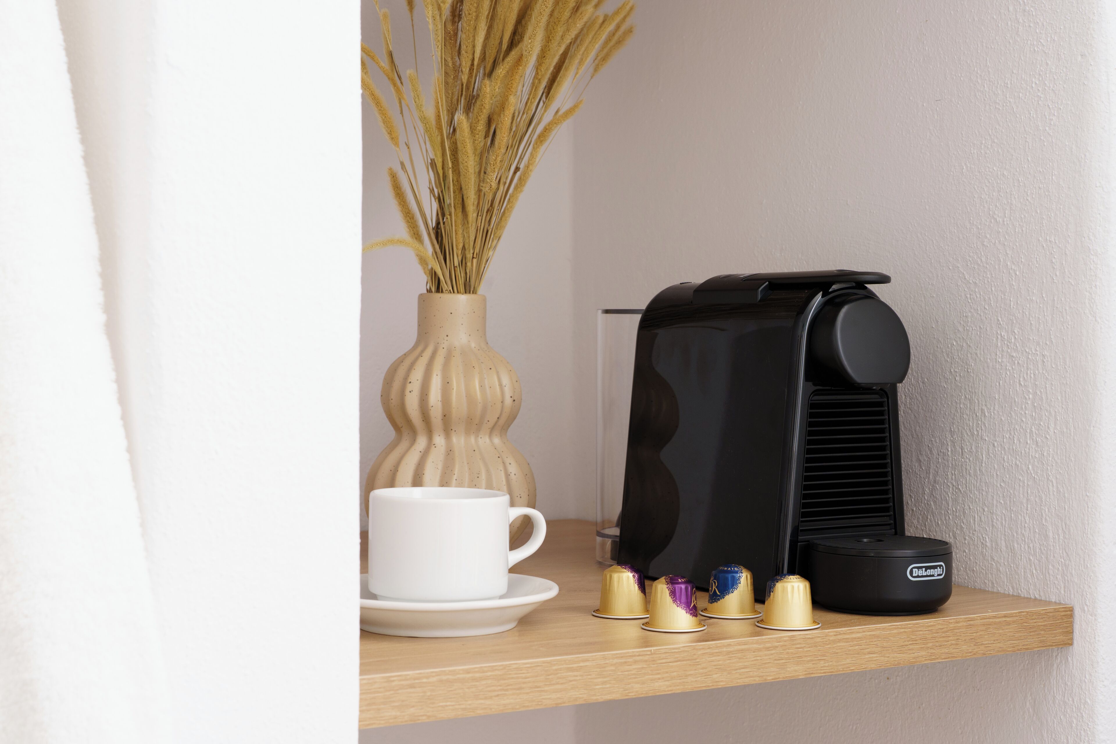 A coffee maker on a shelf with a white cup and a vase of dried plants.