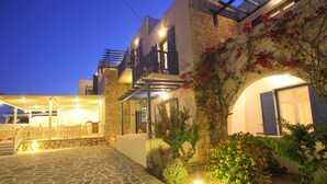 Un patio empedrado con zona de estar cubierta, una escalera que lleva a un balcón y un jardín bien iluminado con plantas en flor.
