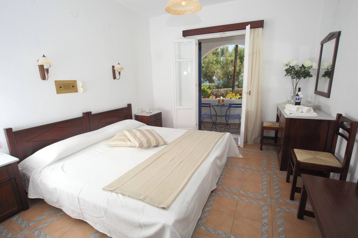 Un dormitorio con cama, mesitas de noche, un escritorio con silla, un espejo y una puerta abierta que da a un balcón con mobiliario exterior.