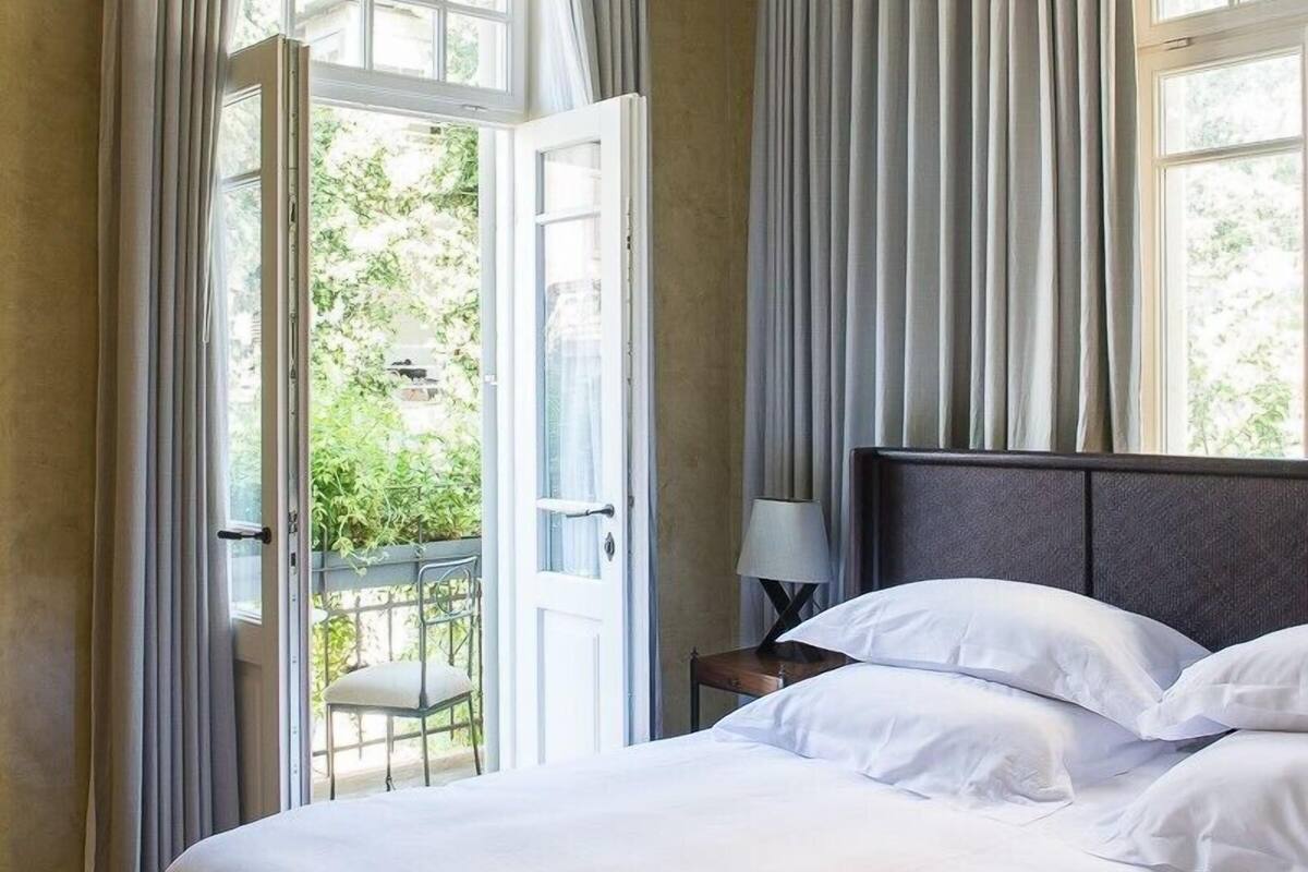 A bedroom with a large bed, a nightstand with a lamp, and a view of greenery through the window.