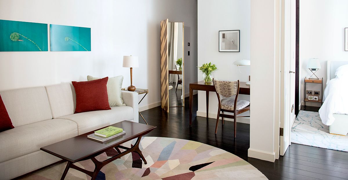 A modern hotel room with a sofa, coffee table, and a dining area with a chair and table.