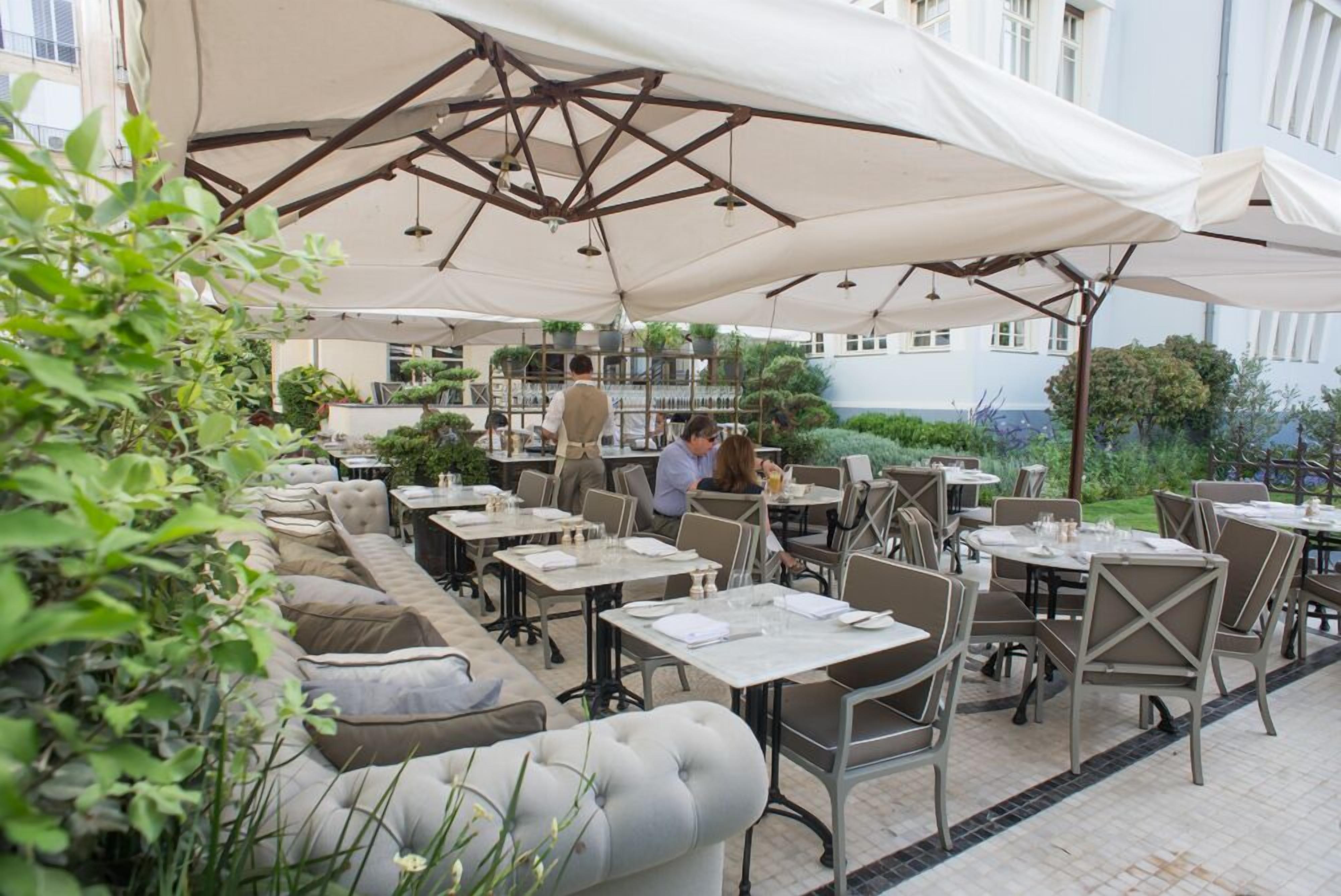 Un área de comedor al aire libre con mesas y sillas, cubierta por sombrillas grandes y rodeada de vegetación.
