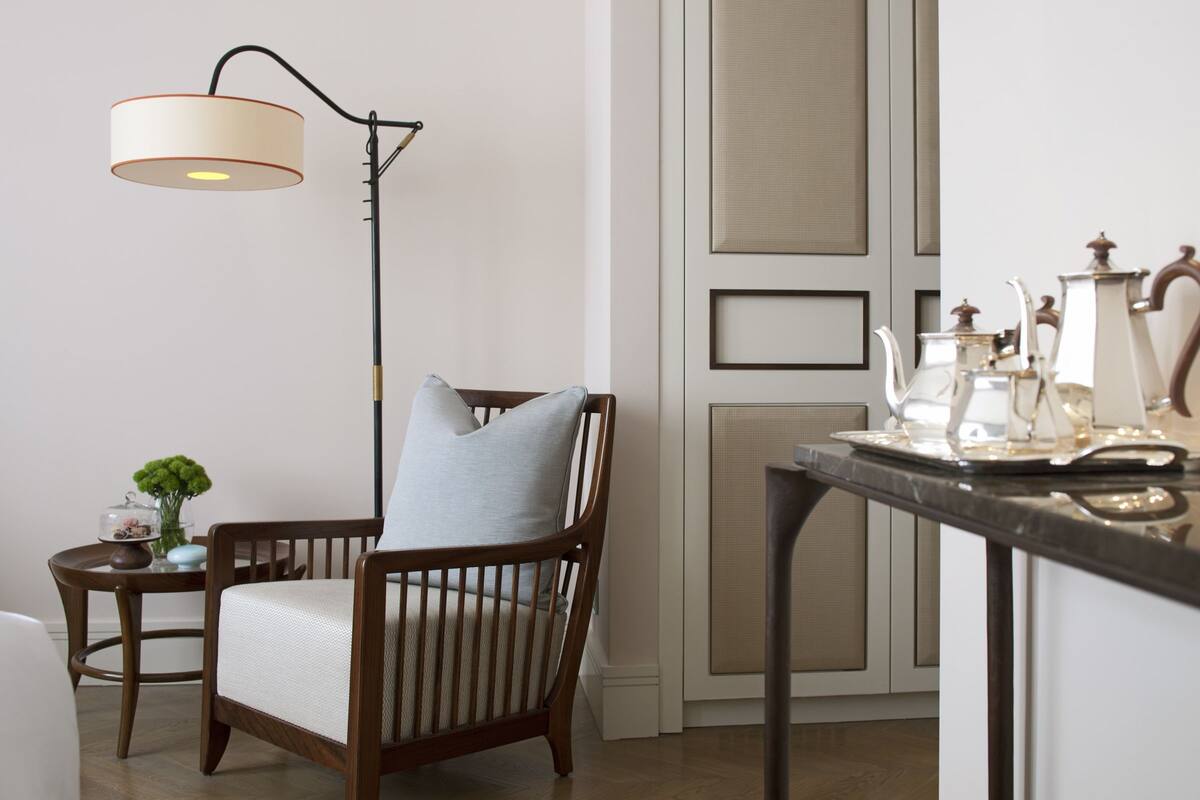 A room with a wooden chair, a small table with a vase of flowers, a floor lamp, and a decorative tray with items on a table.