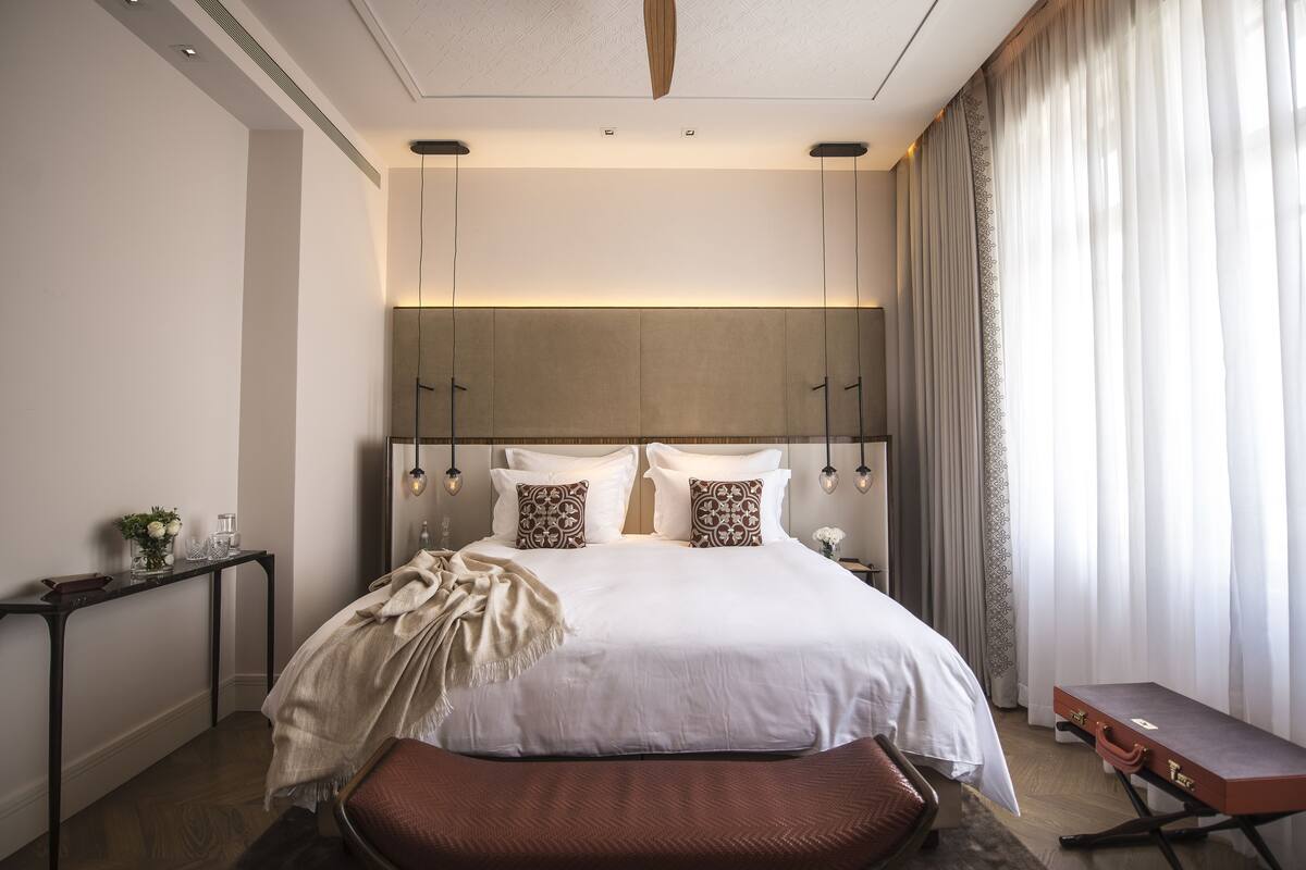 A modern hotel bedroom with a large bed, bedside tables, a bench, and a view of the outdoors.