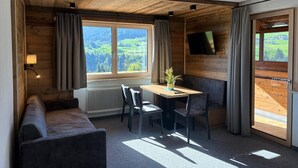 A cozy room with a wooden ceiling, a large window with curtains, a table with chairs, a sofa, a TV mounted on the wall, and a radiator. - Alpenresort Panorama (Oberperfuss)