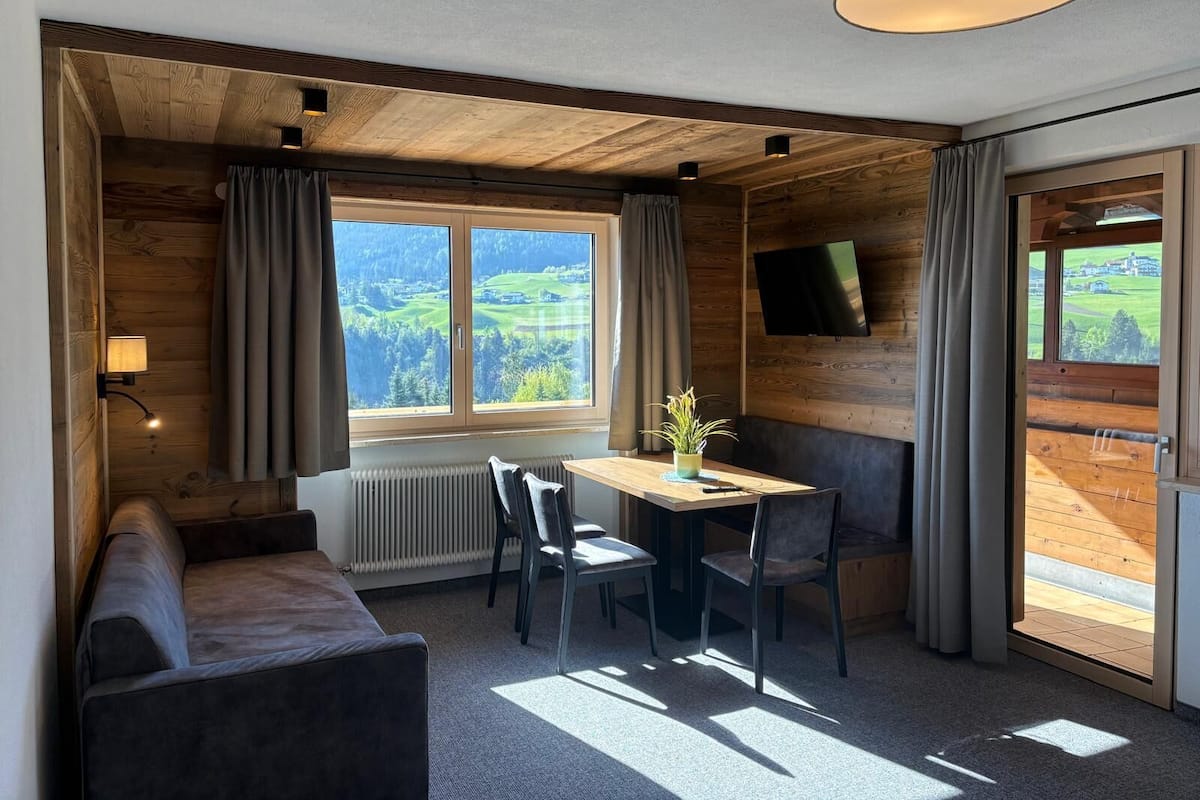 Una habitación acogedora con techo de madera, un ventanal con cortinas, una mesa con sillas, un sofá, un televisor montado en la pared y un radiador.