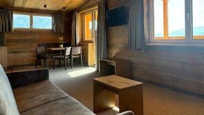A cozy cabin interior with a wooden ceiling, a sofa, a dining table with chairs, and large windows. - Alpenresort Panorama (Oberperfuss)