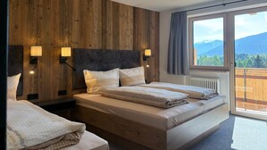 A bedroom with a wooden wall, a large bed, two bedside lamps, and a window with a view of mountains. - Alpenresort Panorama (Oberperfuss)