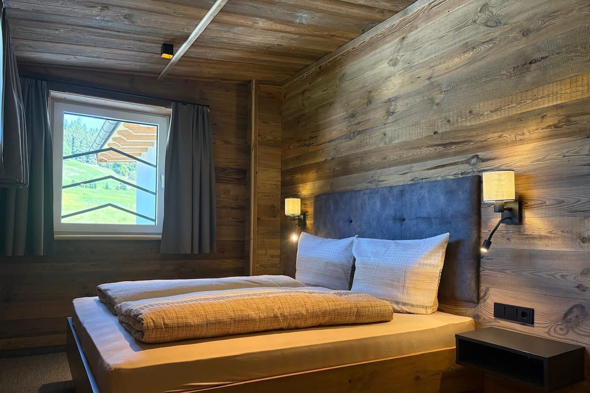 Dormitorio con paneles de madera, una cama, dos lámparas fijadas a la pared y un ventanal con cortinas.