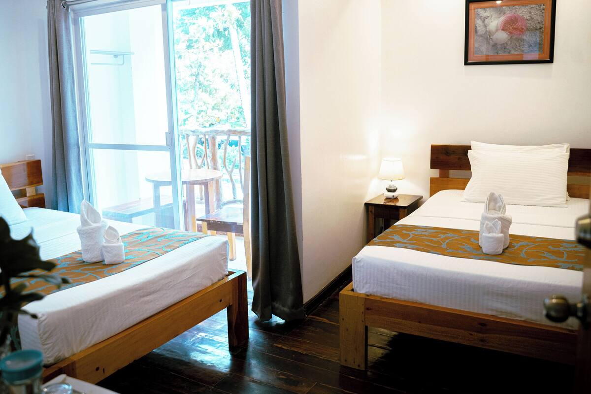 Een hotelkamer met twee bedden, een raam met gordijnen, een klein tafeltje met een lamp en een lijst met een afbeelding aan de muur.