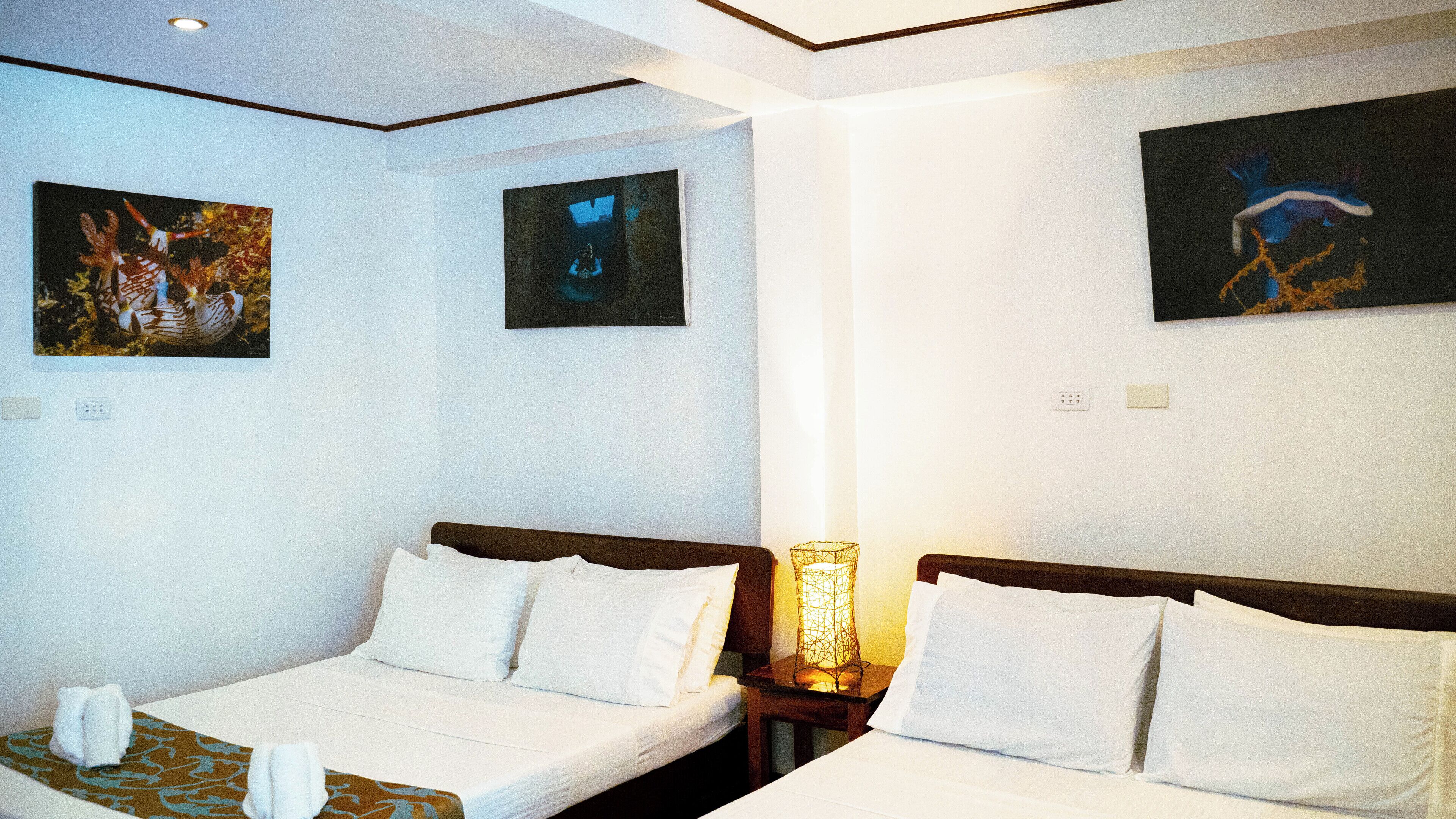 A hotel room with two single beds, a nightstand with a lamp, and two paintings on the walls.