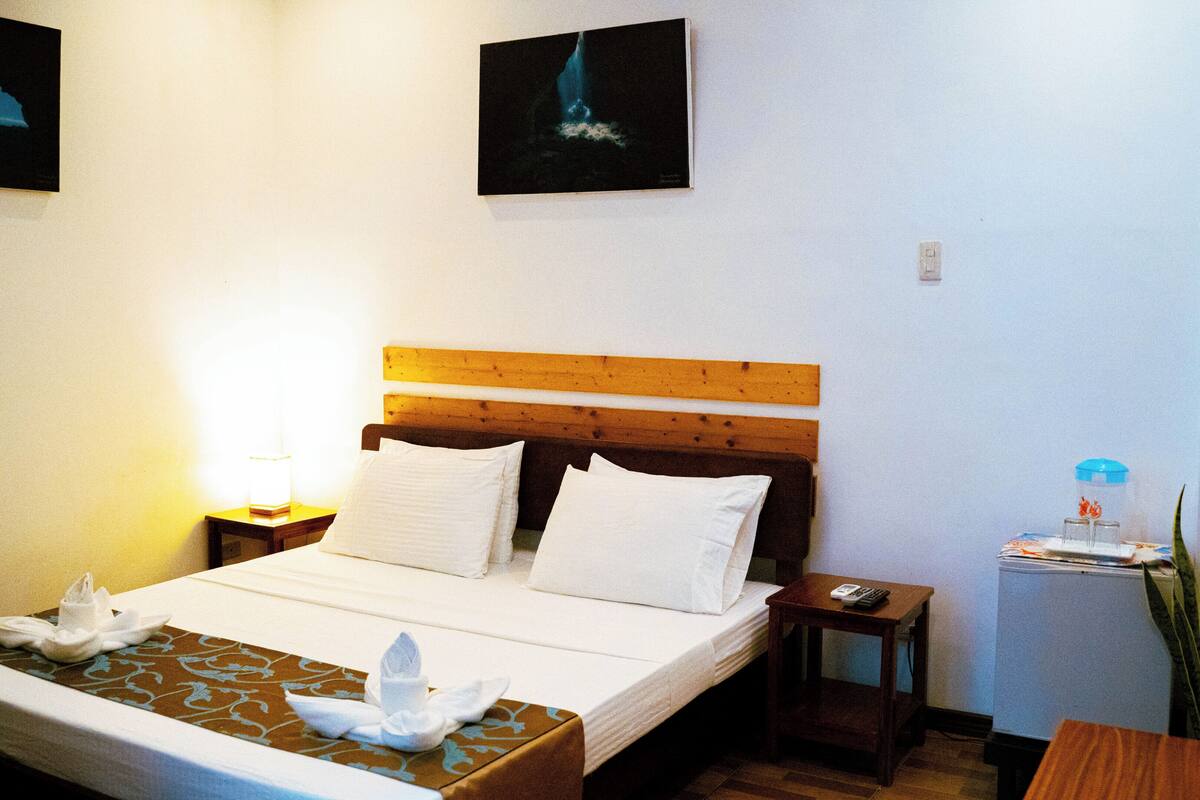 Een hotelkamer met een houten hoofdeinde, een bed met witte lakens, een nachtkastje met een lamp en een tafeltje met een vaas en een afstandsbediening.