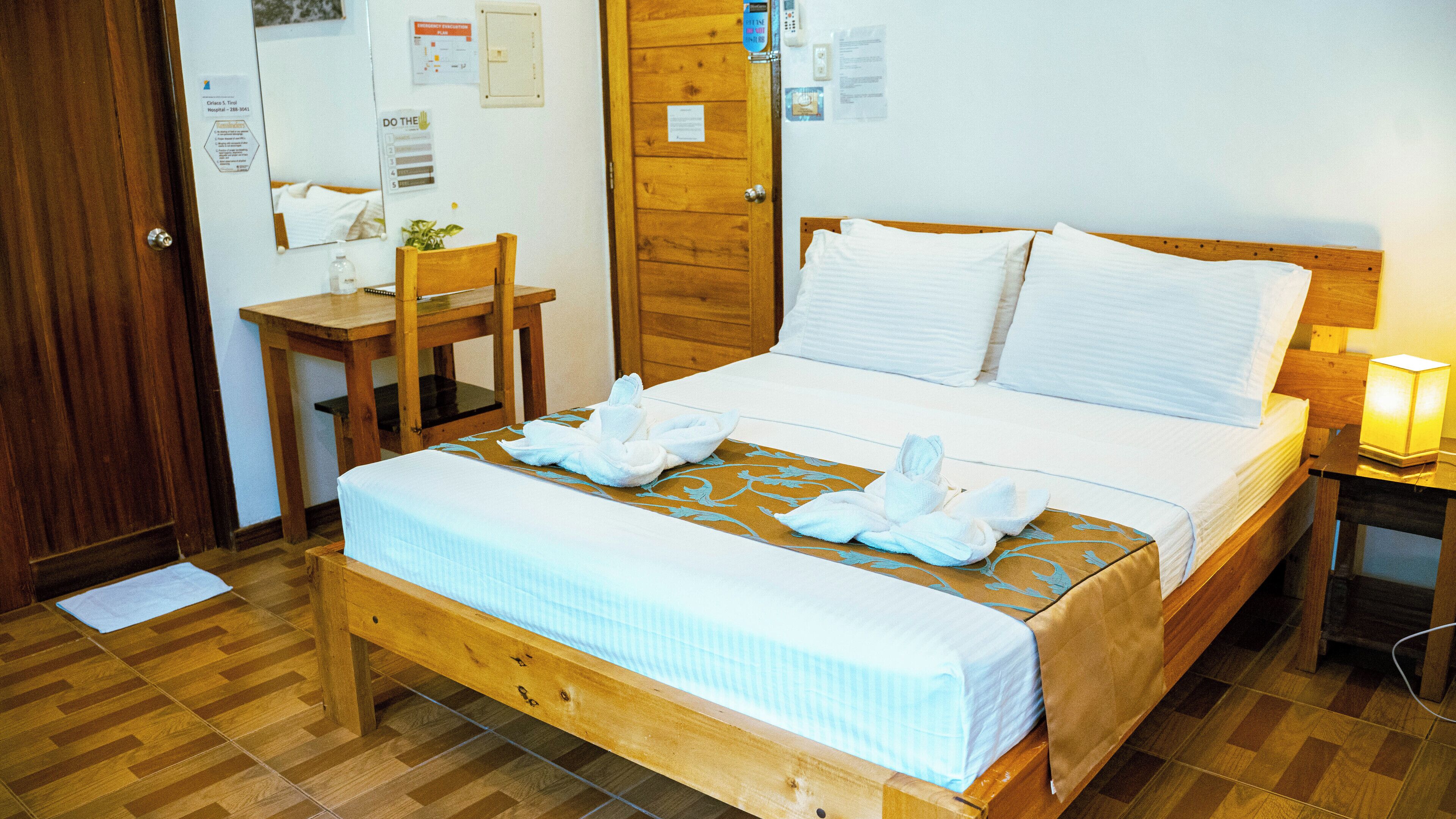 Habitación de hotel con una cama de madera, una mesita de noche con lámpara, una silla de madera y un escritorio con computadora.