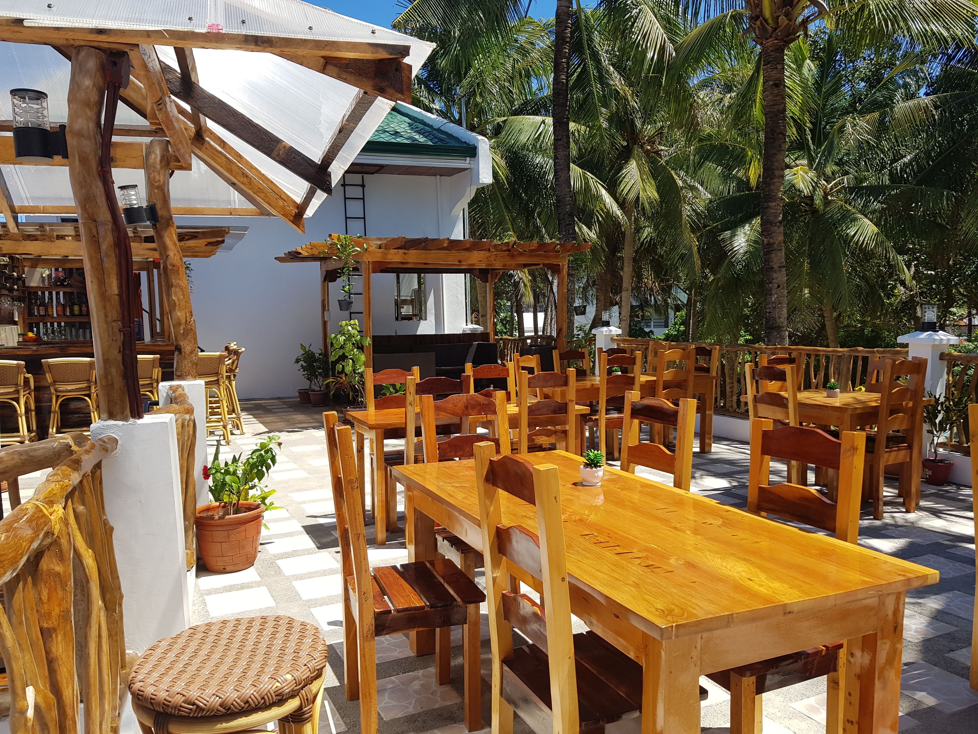 An outdoor patio area with wooden tables and chairs, surrounded by potted plants and palm trees.