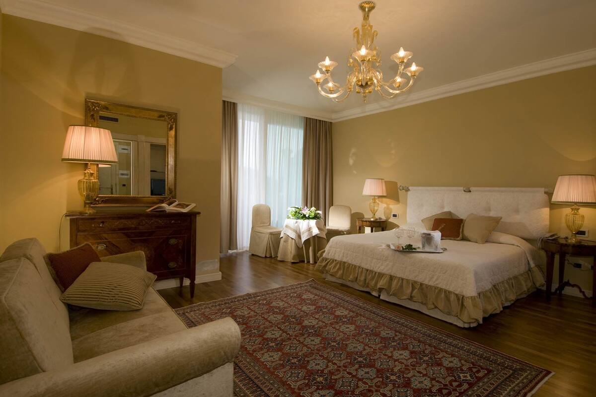 A hotel room with a large bed, a sitting area with a chair and a small table, a dresser, a mirror, and a chandelier.
