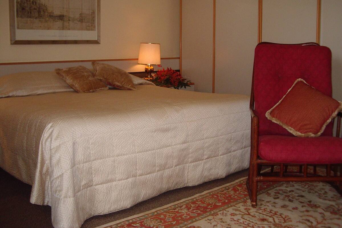 A hotel room with a bed, pillows, a nightstand with a lamp, a chair, and a framed picture on the wall.