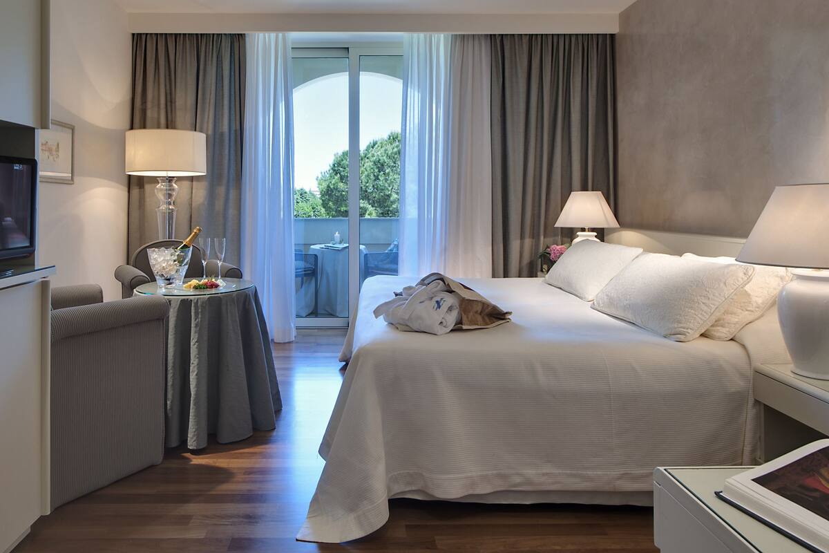 A hotel room with a bed, a TV, a table with a bottle of champagne, and a balcony with a table and chairs.