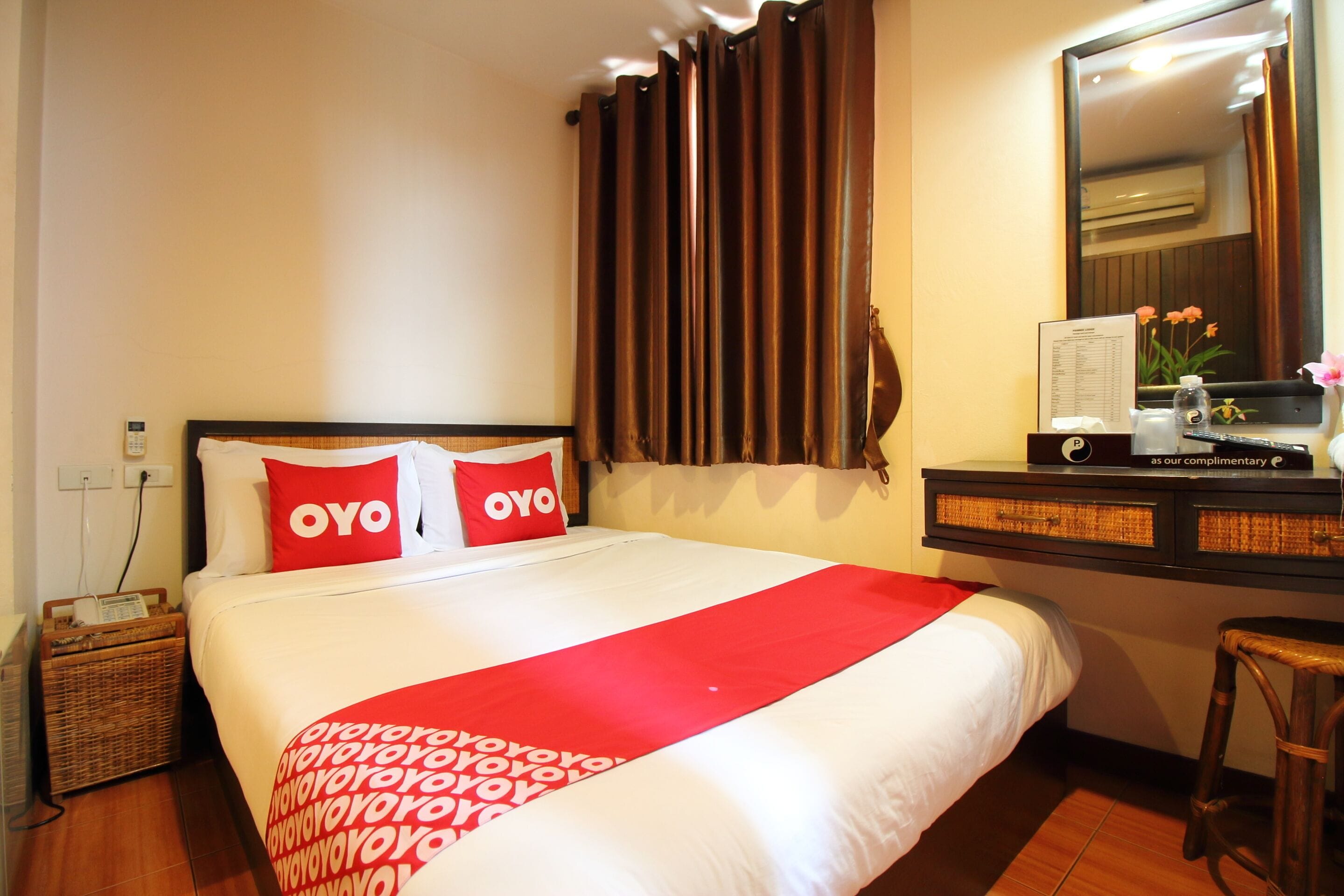 Camera d'albergo con un letto grande, due cuscini rossi con la scritta "OYO", una coperta in stile rosso, un comodino in legno con una lampada, un tavolino con un telecomando e un vaso con fiori, e uno specchio sopra il letto.