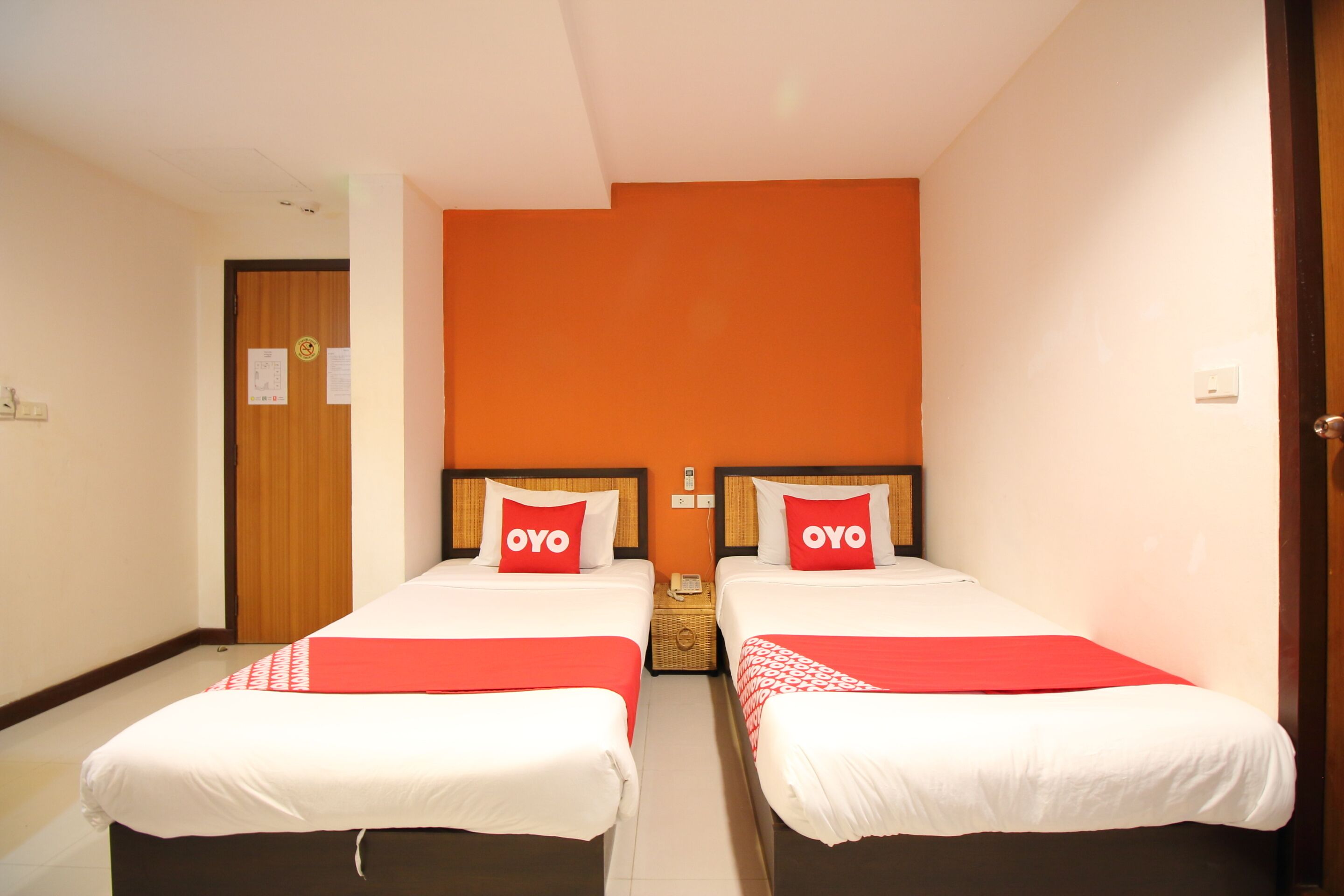 A hotel room with two beds, each with a red pillow labeled "OYO," white bedding, and a red throw blanket.