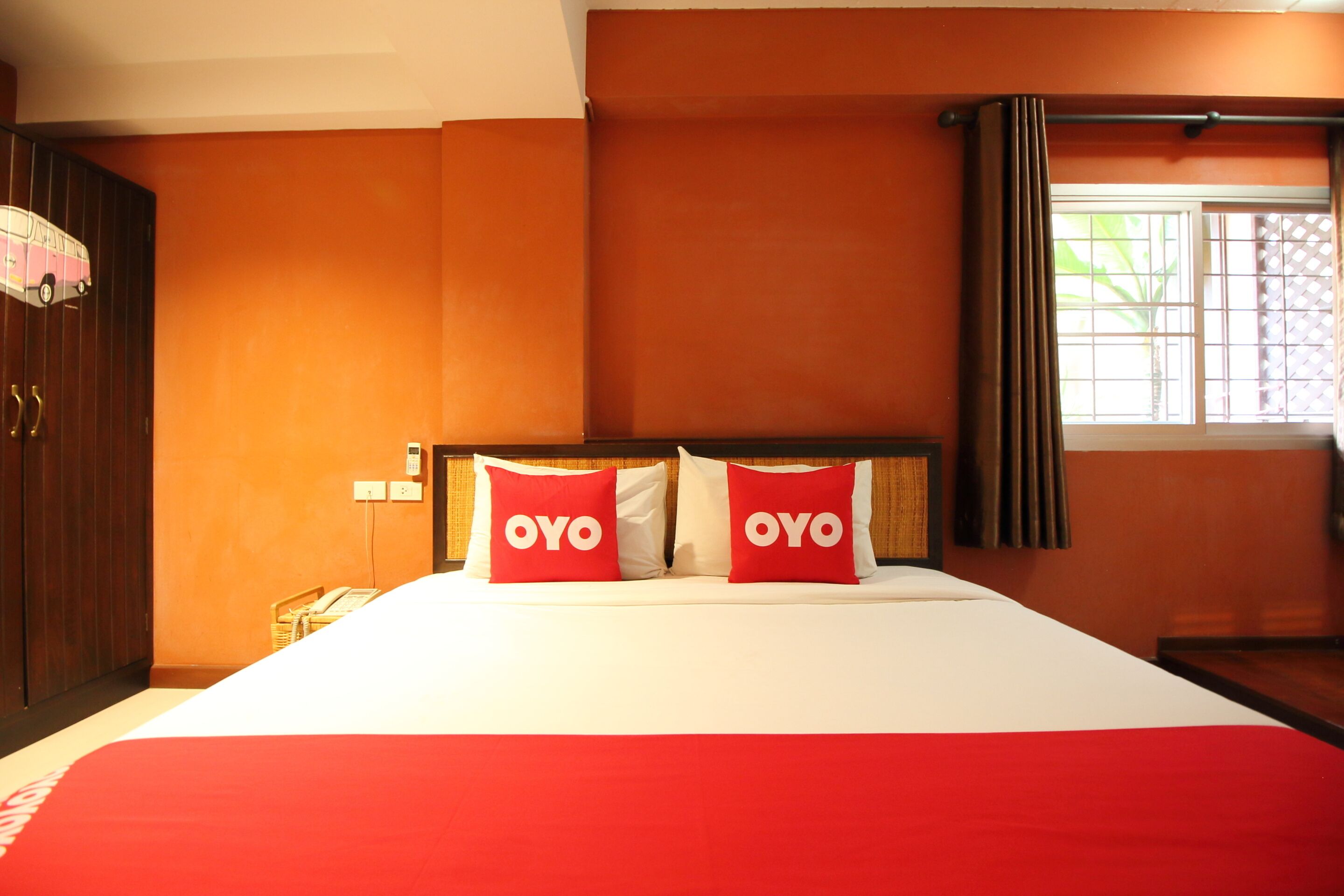 A hotel room with a large bed, two red pillows with 'OYO' branding, a wooden wardrobe, and a window with a view of greenery.