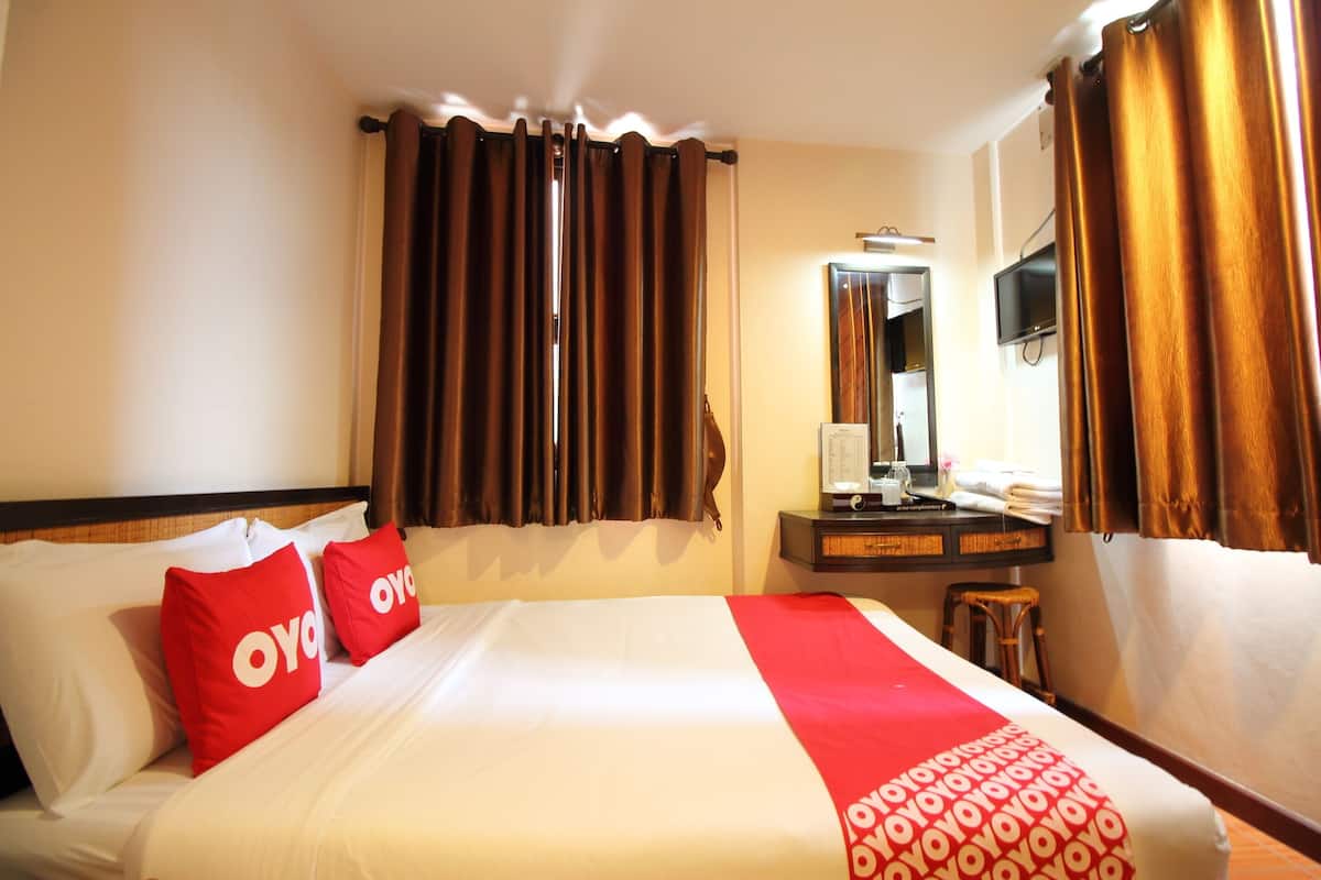 Een hotelkamer met een groot bed, twee rode kussens, een rood-witte sprei, een houten nachtkastje, een spiegel en bruine gordijnen.