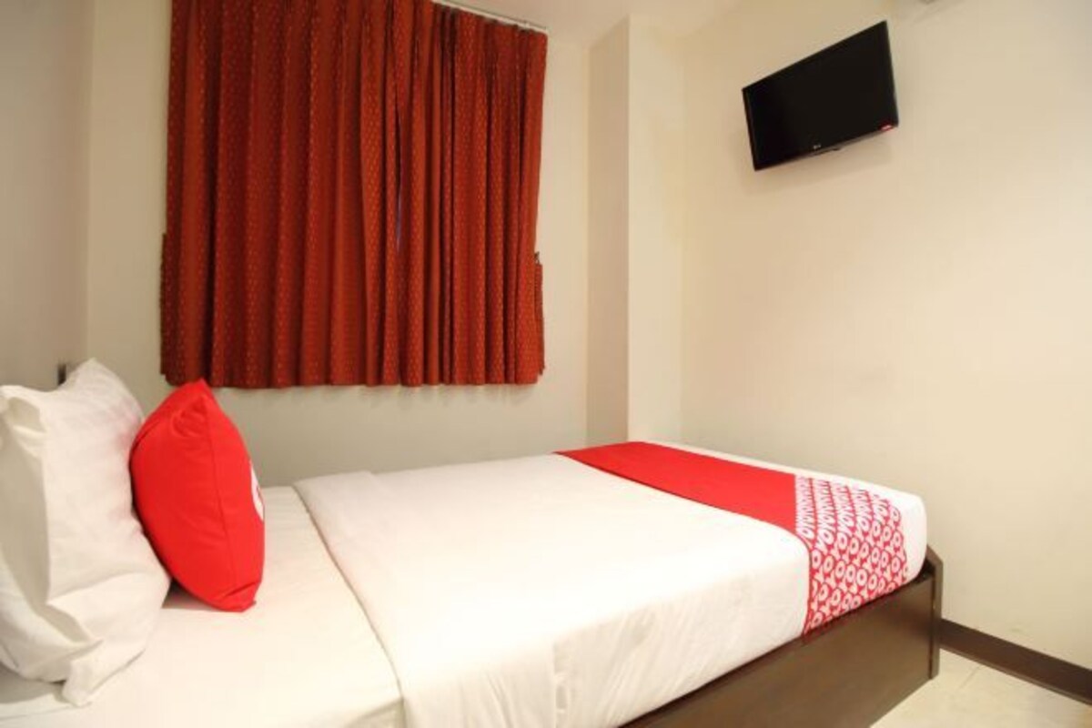 Una cama individual con ropa de cama blanca y una almohada decorativa roja, una manta roja y un televisor montado en la pared.