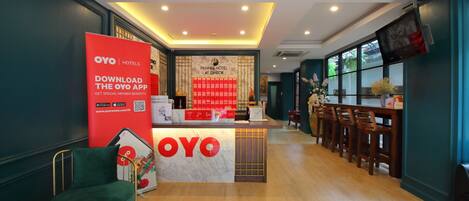 A hotel reception area with a reception desk, a red OYO app advertisement, and a bar area with wooden chairs.