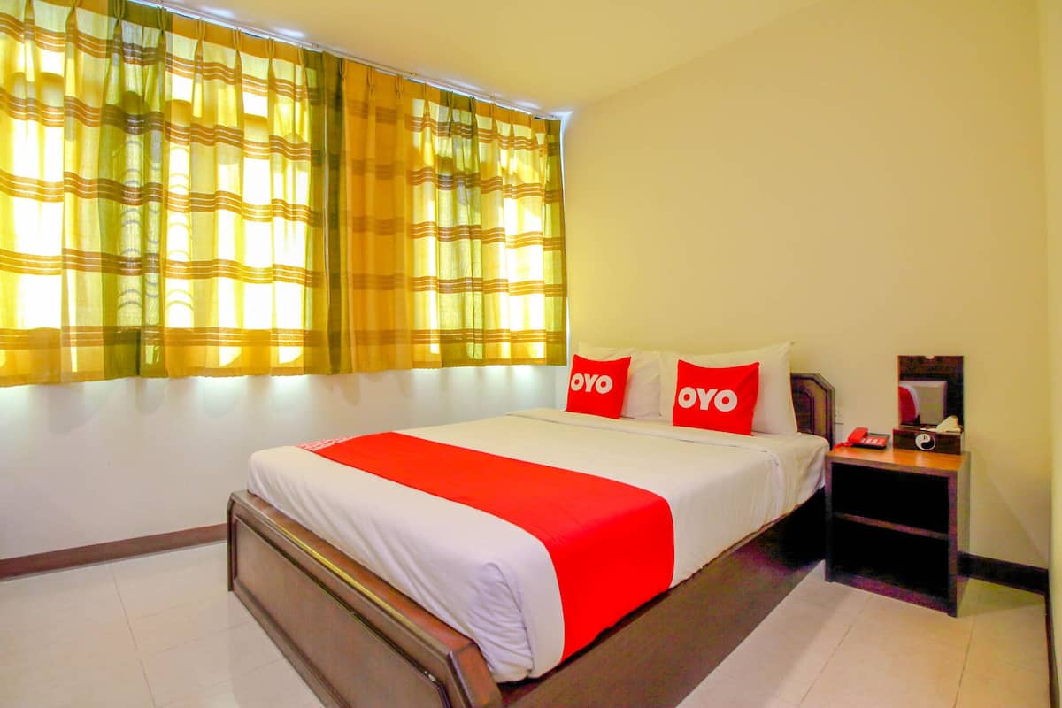 A hotel room with a bed, two red pillows, a bedside table with a phone, and a window with yellow curtains.