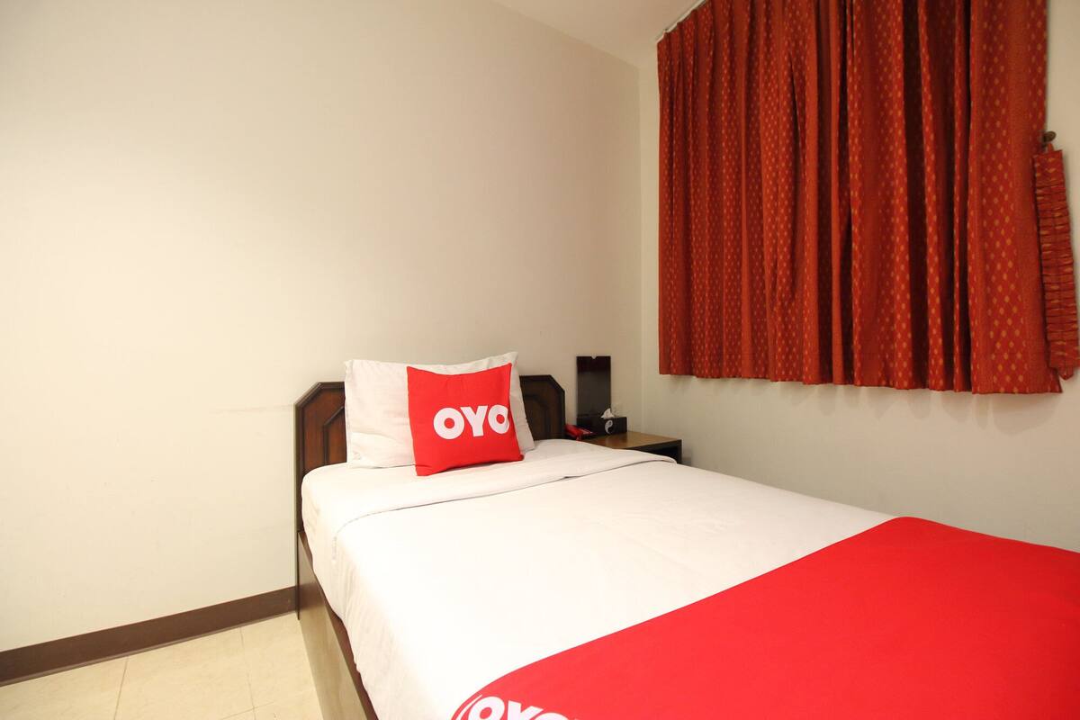 A hotel room with a bed, a red and white pillow, a red blanket, a nightstand, a telephone, and a window with red curtains.