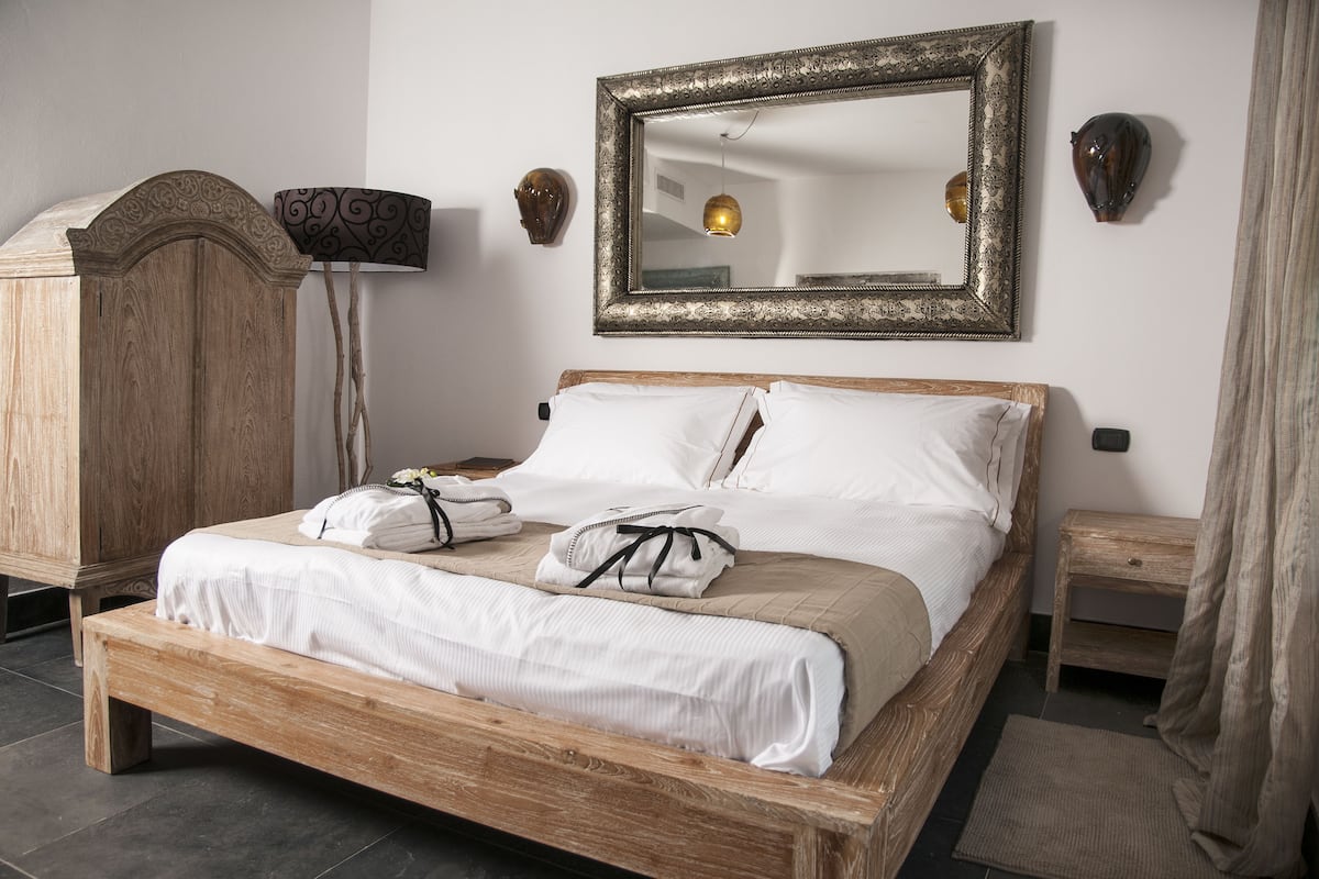 A wooden bed with white bedding, a large ornate mirror, and a lamp on a stand.