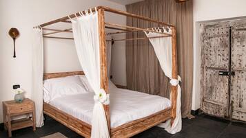 Un lit à baldaquin avec du linge de lit blanc et des rideaux, une table de chevet en bois ornée d’un vase, et une porte rustique.