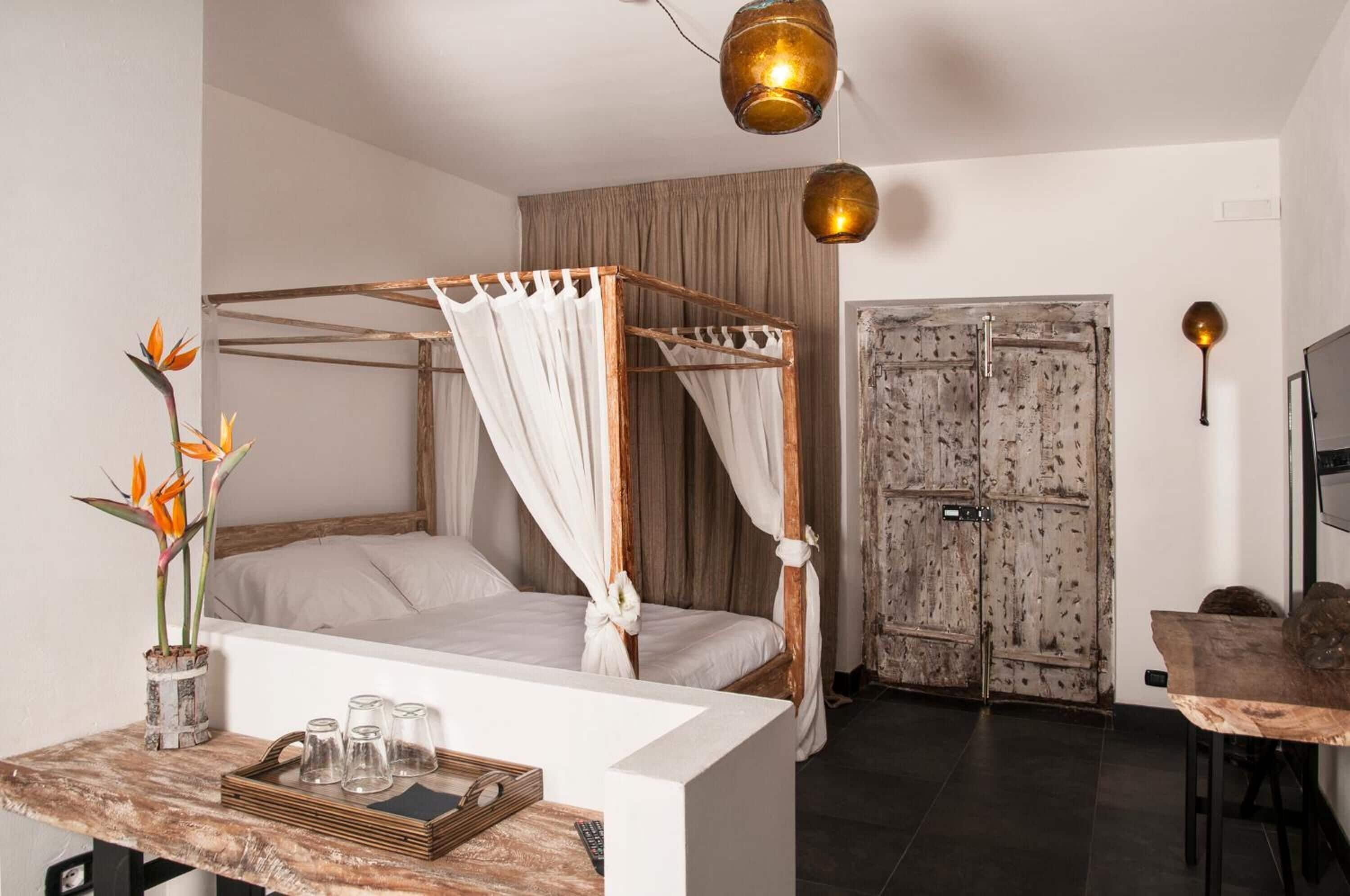Ein Bett mit Baldachin, ausgestattet mit weißen Leinen, ein Nachttisch aus Holz mit einem Tablett mit Gläsern und eine Vase mit Blumen.