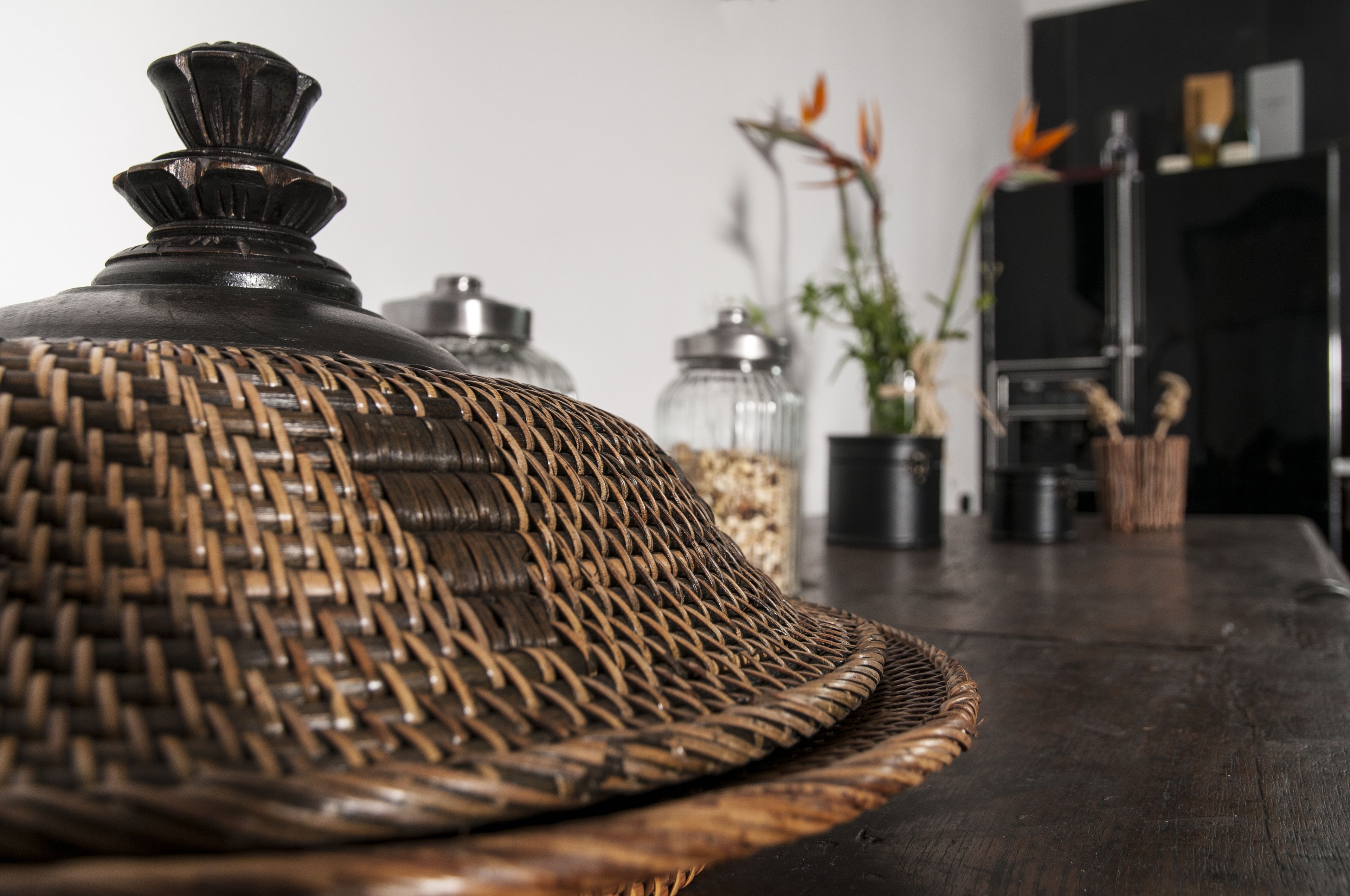 Una cesta de mimbre con tapa sobre una mesa de madera, frascos de vidrio y una planta en maceta al fondo.
