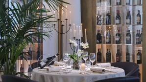 A well-lit dining area with a round table set for two, surrounded by dark chairs. A large potted plant is to the left, and a wine rack with bottles is in the background. - Europa Terme (Abano Terme)