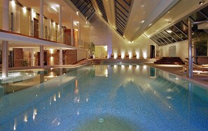 Una piscina interior con agua cristalina, rodeada por un diseño arquitectónico moderno con barandillas de vidrio y una escalera que lleva a un nivel superior.