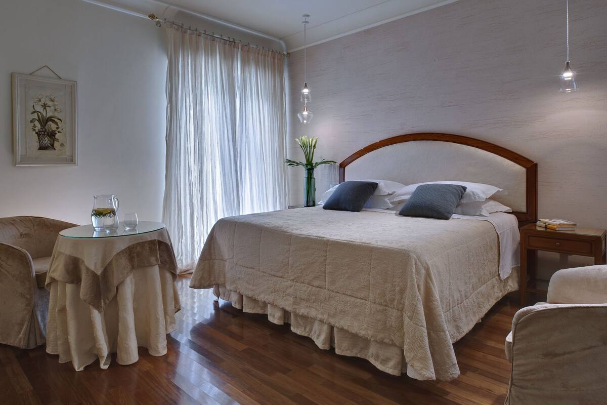 Un dormitorio ordenado con una cama grande, una mesita redonda con una jofaina y vasos, dos sillas, un florero con flores y un cuadro en la pared.