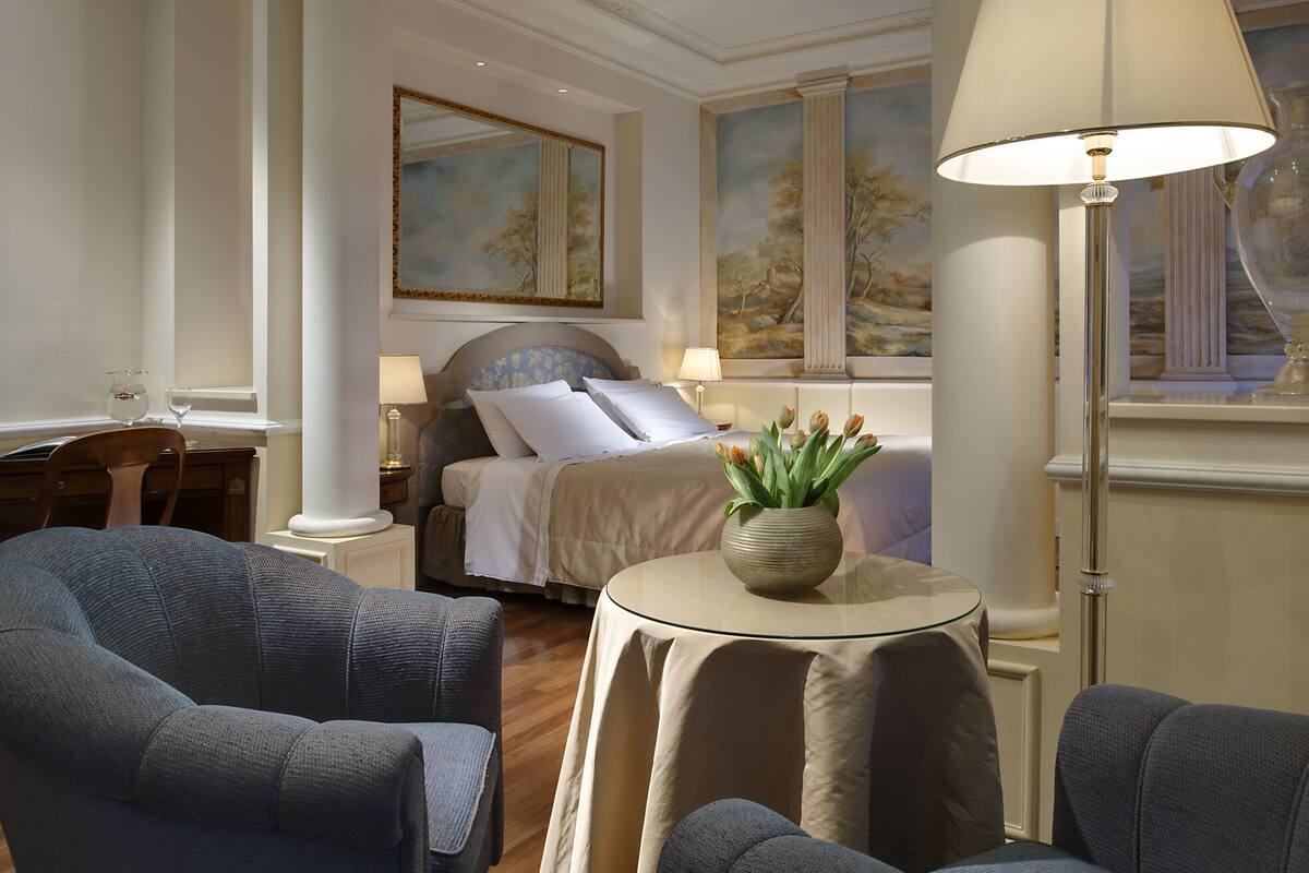 Habitación de hotel con una cama, dos sillones, una mesita con un jarrón de flores y una lámpara.