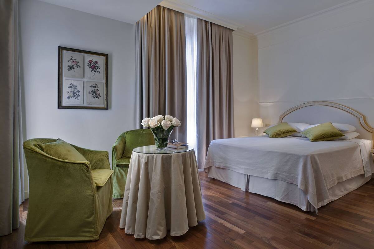 Habitación de hotel con cama, dos sillas, una mesita con un florero y grabados botánicos en la pared.