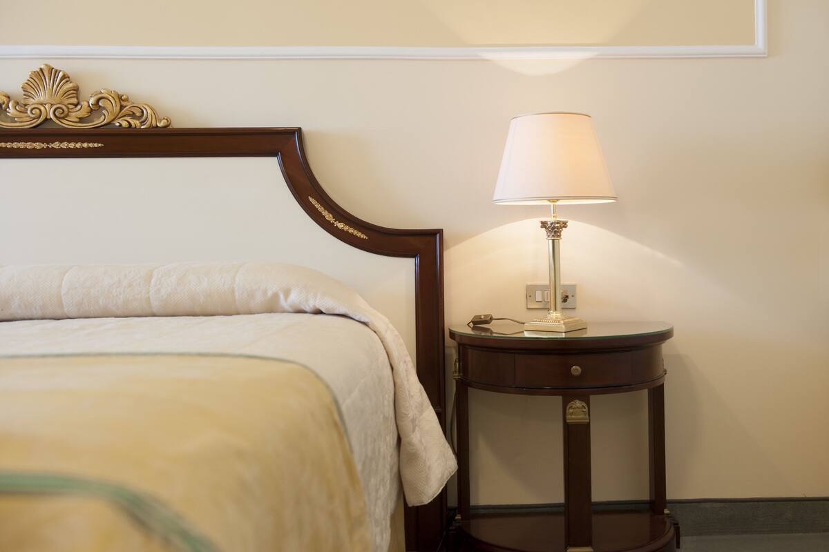 Una cama bien hecha con cabecera, una mesita de noche con lámpara y un interruptor de luz en la pared.