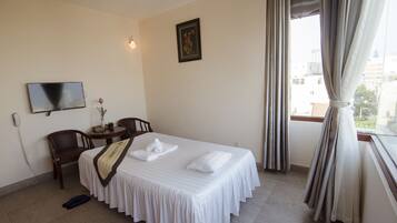 Habitación de hotel con cama, una silla, una mesita, televisor y ventana con cortinas.