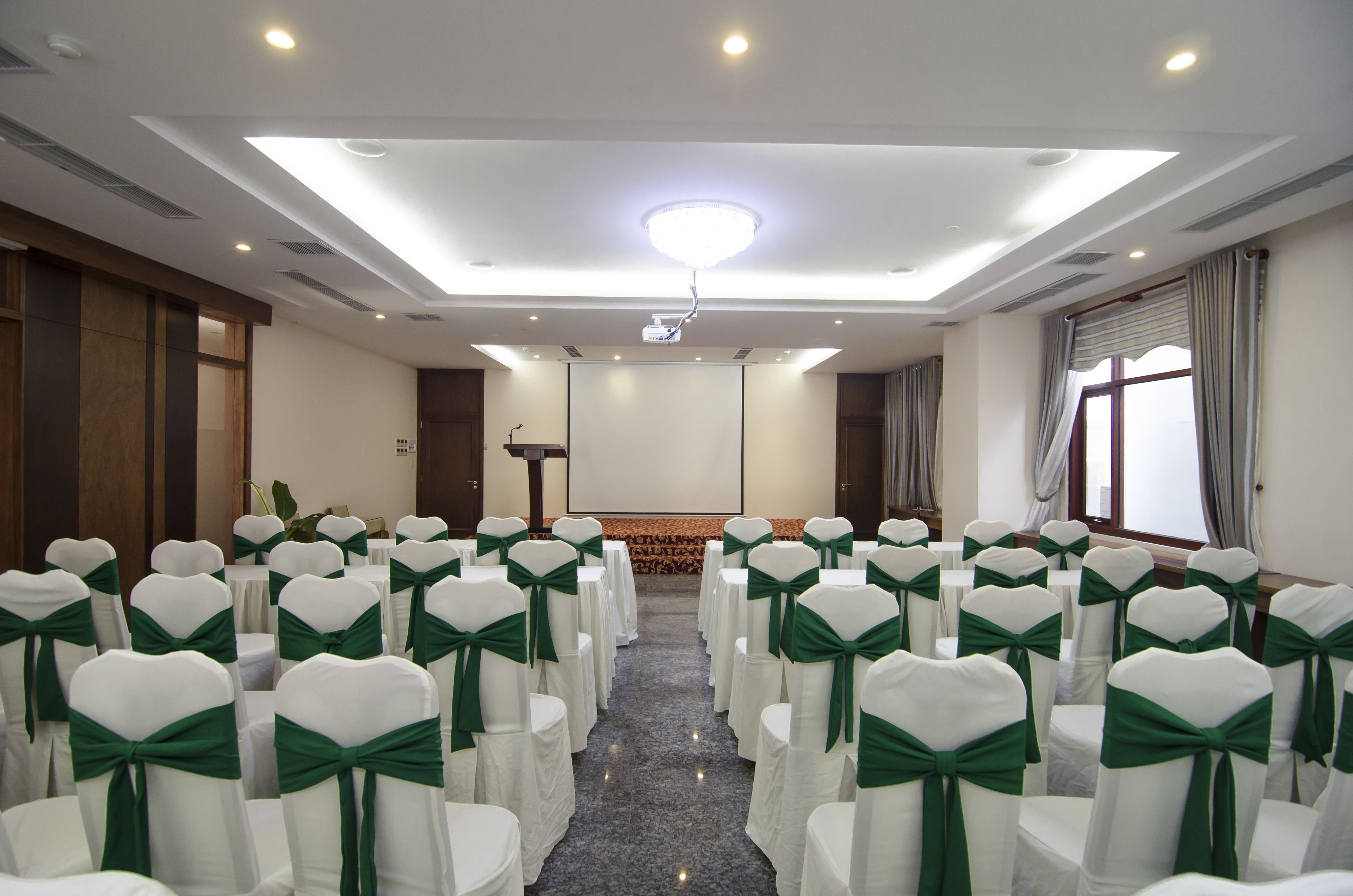 Une salle de conférence avec des rangées de chaises blanches ornées de rubans verts, un pupitre, un projecteur et un grand écran.