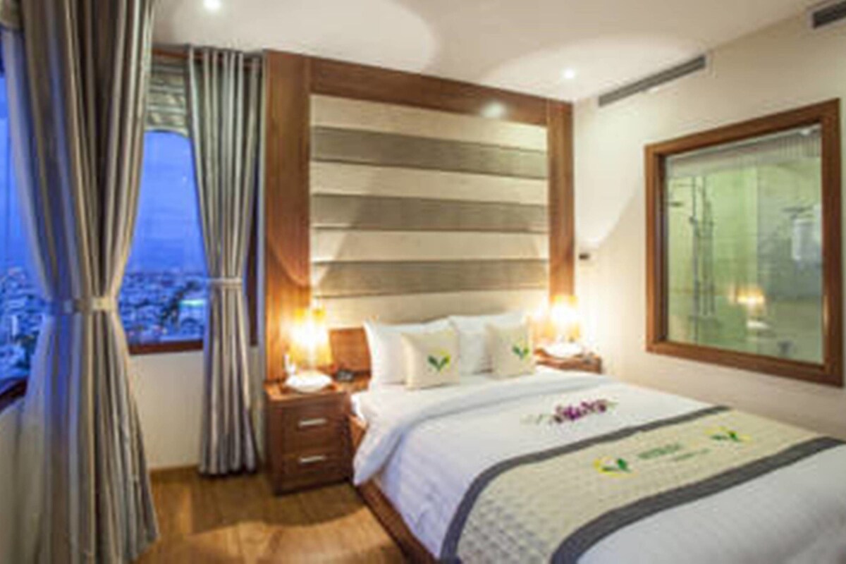 A hotel room with a large bed, wooden headboard, nightstands, and a city view through the window.