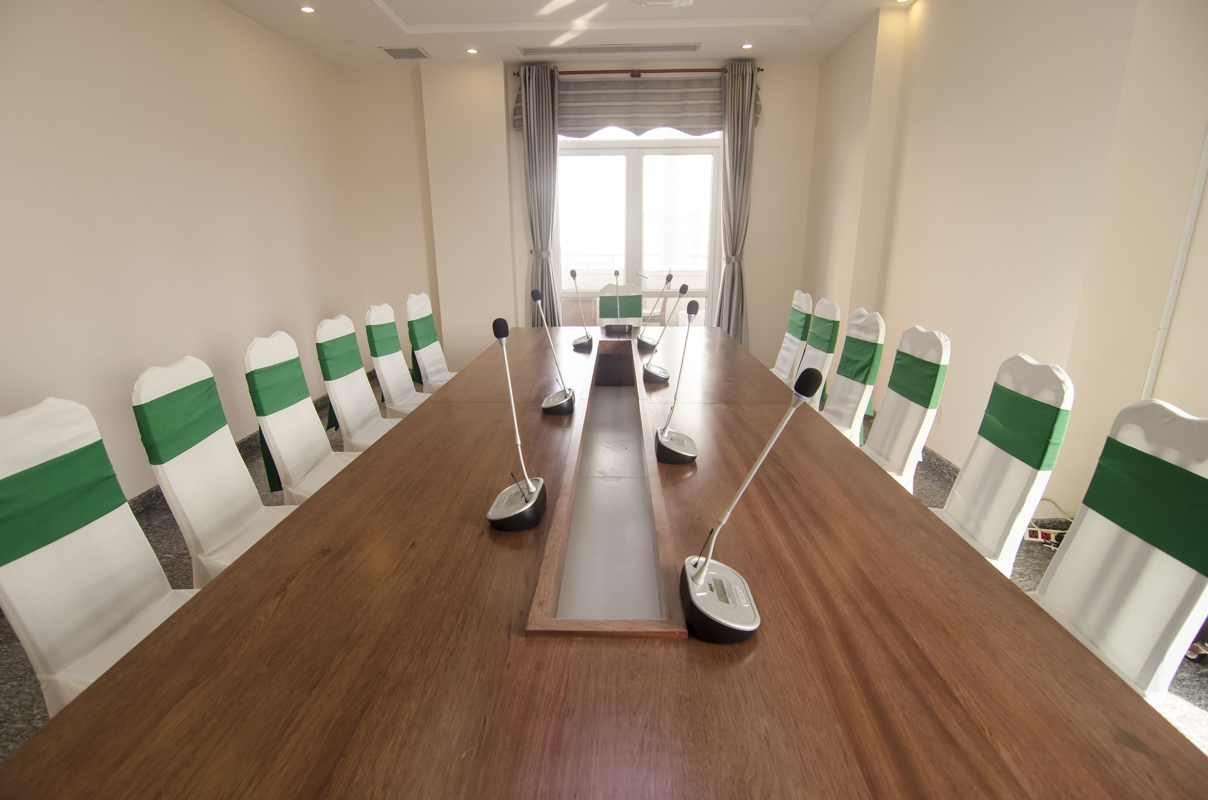 A conference room with a long table, chairs with green sashes, microphones, and a large window with curtains.