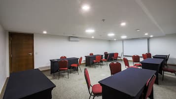 Uma sala de conferência com mesas, cadeiras, tela de projetor e um aparelho de ar condicionado.