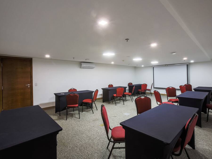 Uma sala de conferência com mesas, cadeiras, tela de projetor e um aparelho de ar condicionado.