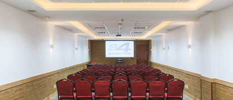 Un salón de conferencias con hileras de sillas rojas, una pantalla de proyector mostrando "EVENTOS" y un revestimiento de madera en la pared.